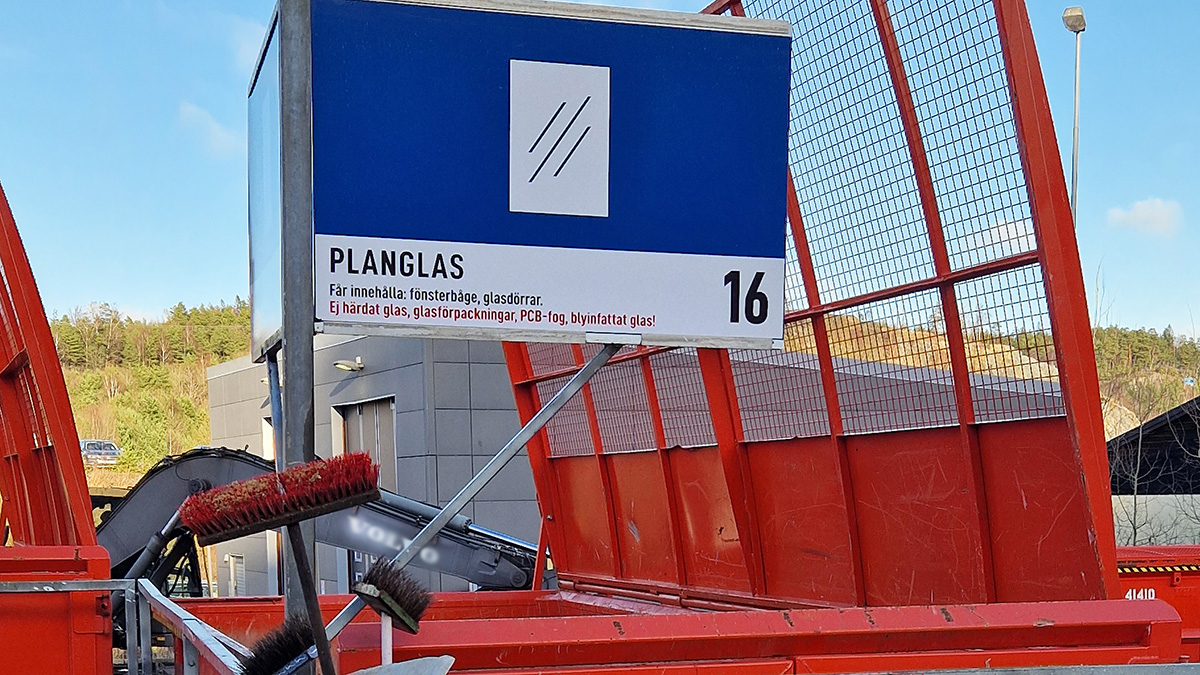 En orangefärgad container på återvinningscentralen. Ovanför sitter en skylt. På skylten står det "Planglas". 