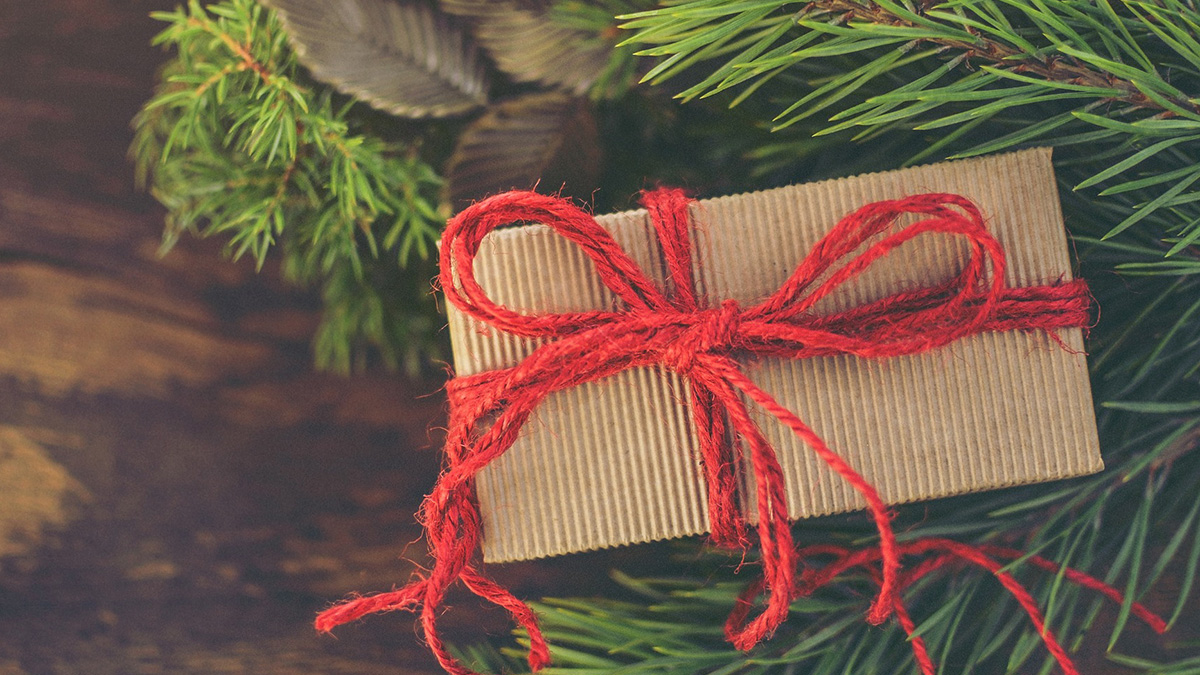 En liten julklapp inslagen i ljusbrunfärgat papper med ett rött band av garn runt, ligger intill granris. 