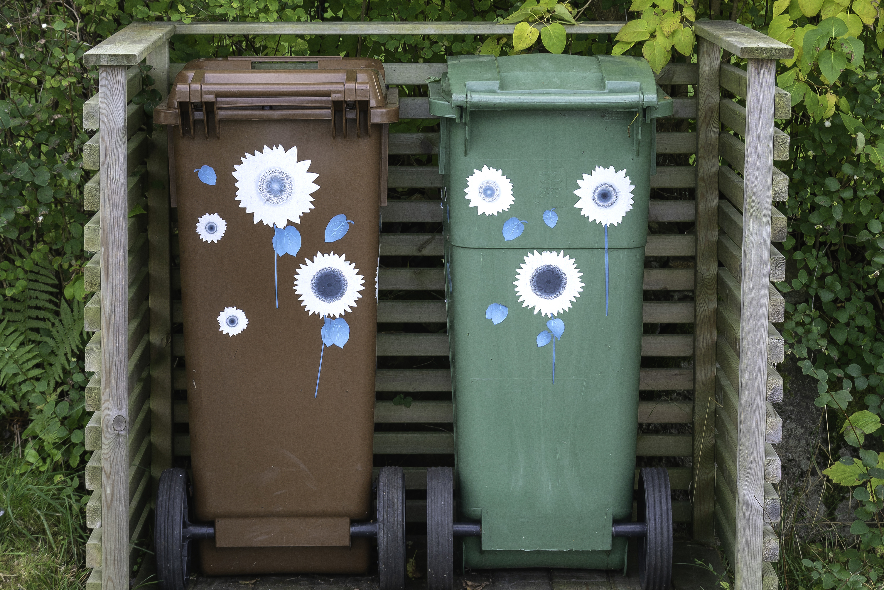 Två soptunnor; en grön och en brunfärgad. Båda står utomhus och har klistermärken i form av blommor på. 