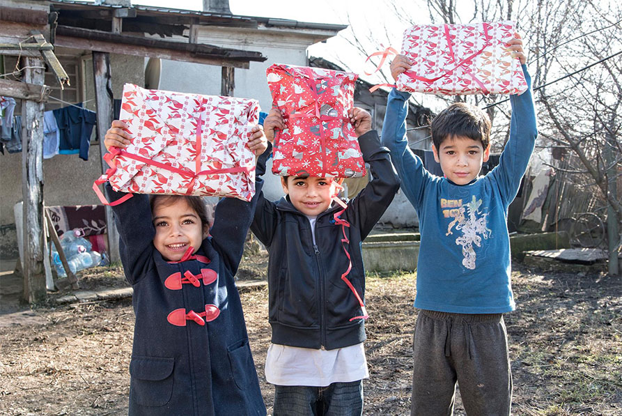 Tre barn håller upp varsin julklapp och ser glada ut. Bakom dem skymtasett fallfärdigt hus. 