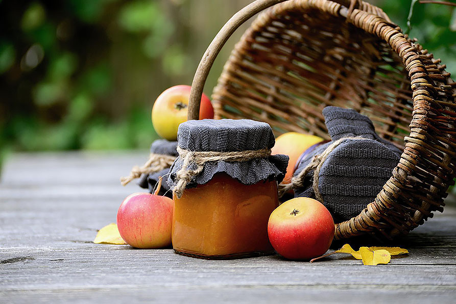 En korg med några äpplen i. Bredvid står glasburkar med hemmagjort äppelmos. 