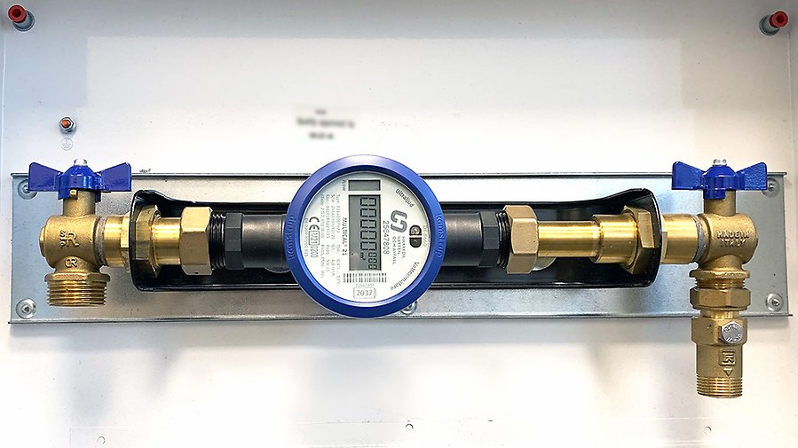 En vattenmätare sitter fast i ett väggfäste (konsol) som håller mätaren och rören med avstängningsventiler på plats. 