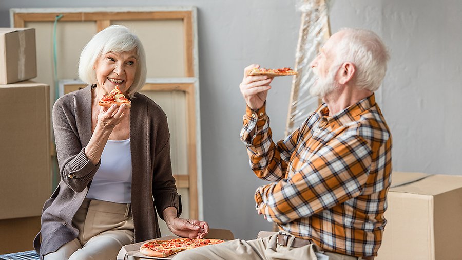 En man och en kvinna sitter bland flyttkartonger och möbler. De skrattar och äter pizza. 