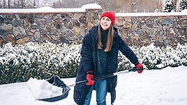 En ung kvinna skottar snö. Hon har långt ljusbrunt hår och är klädd i en lång täckjacka i mörkblått. På huvudet har hon en stickad röd mössa. Kvinnan ser glad ut. 